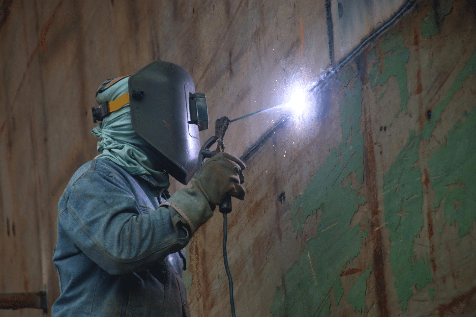 Welding worker in protective helmet and gloves welds a metal seam, bright arc lighting the wall.