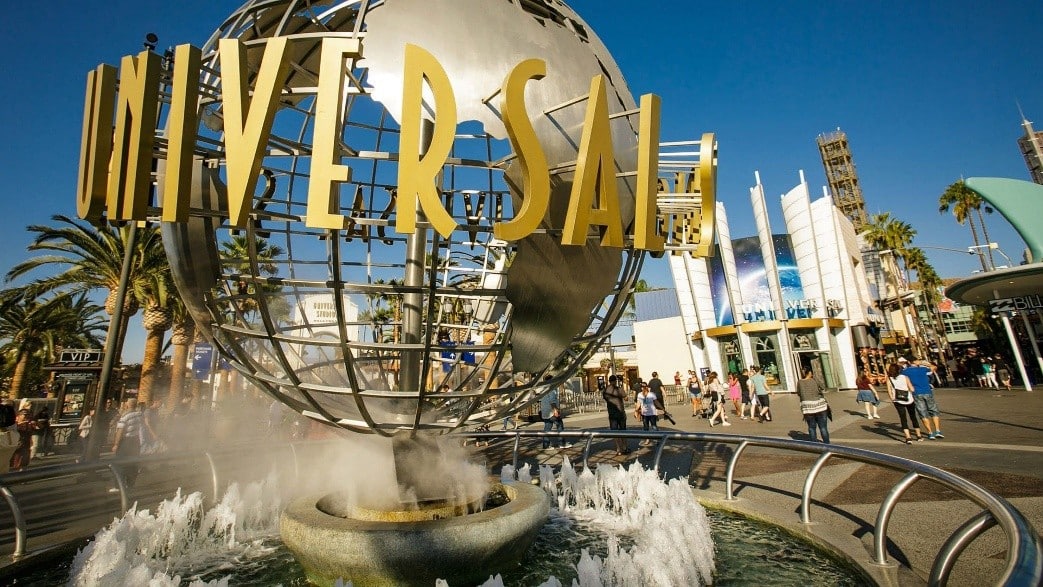 Iconic Universal Studios globe sculpture with a tall gold 'UNIVERSAL' sign above a fountain at the park entrance, palm trees in the background.