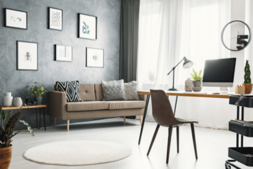 Bright contemporary living room with a beige sofa, wall art, and a wooden desk featuring an iMac and a desk lamp under natural light.