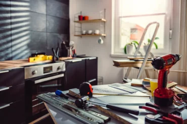 Renovation workspace with a red cordless drill and assorted tools on a cluttered workbench, wood planks and a ladder by a bright window.