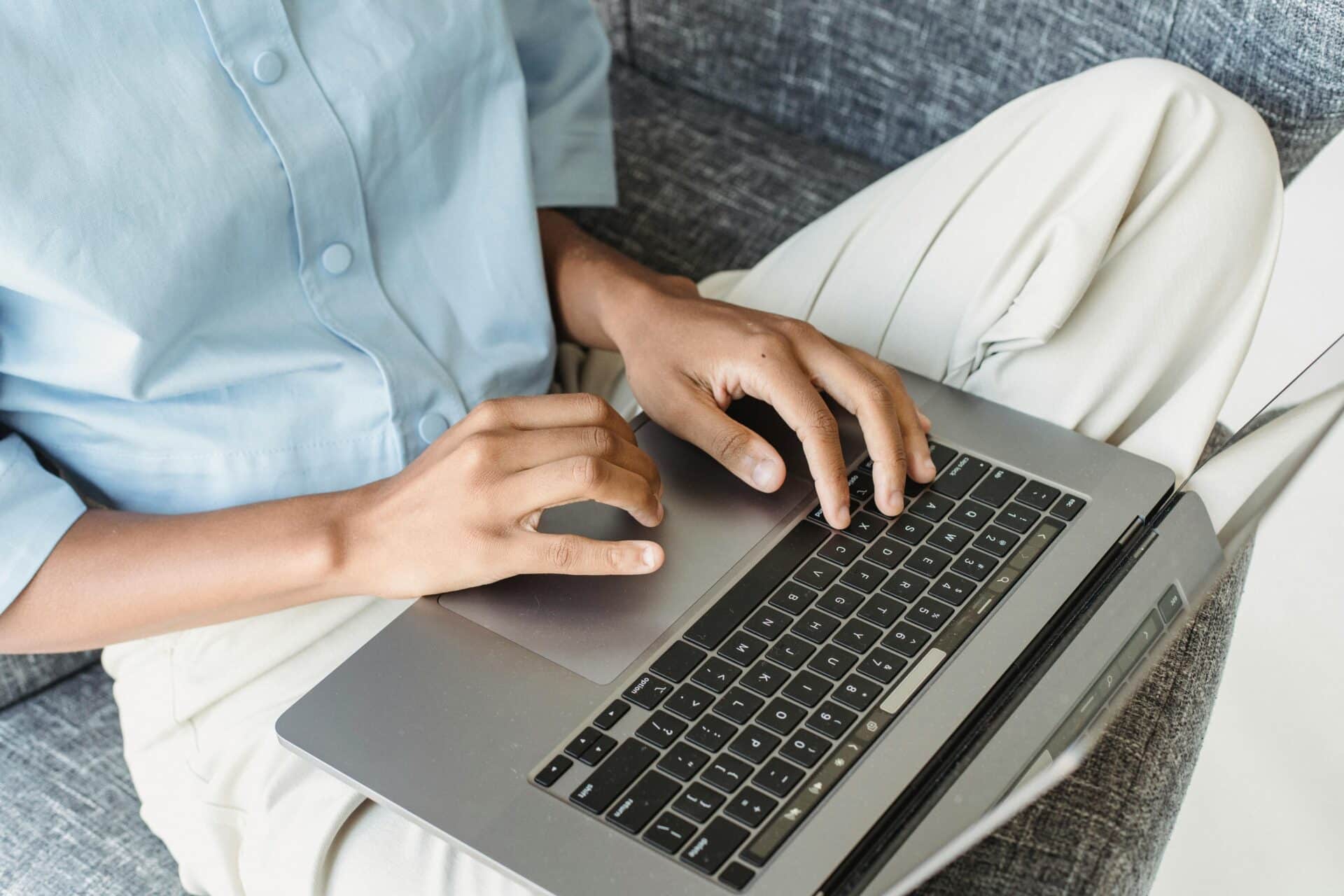 Person wearing a light blue shirt typing on a laptop resting on their lap on a couch.