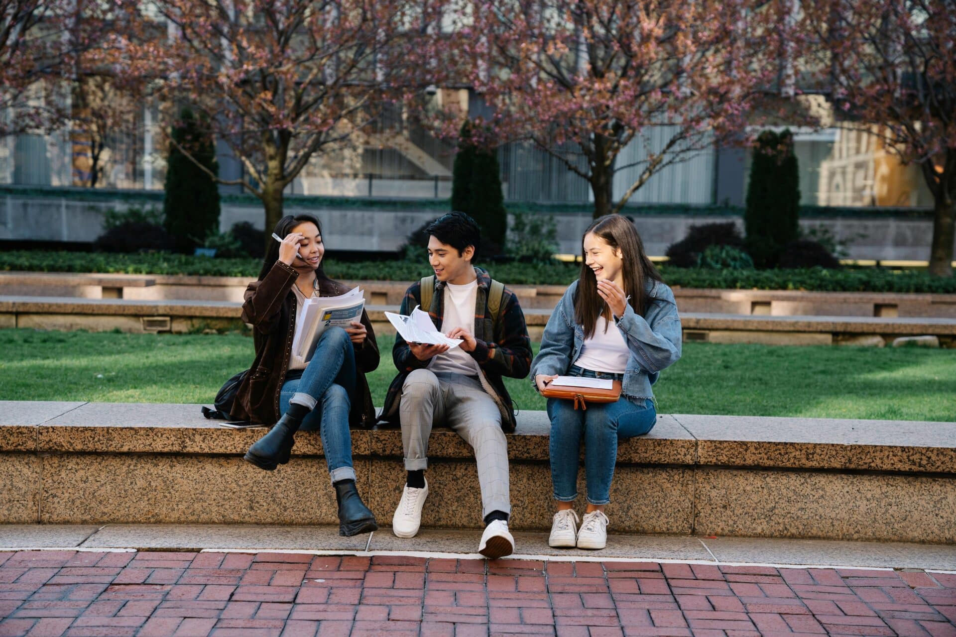 Three students sit on stone steps in a park, chatting and looking at papers together.