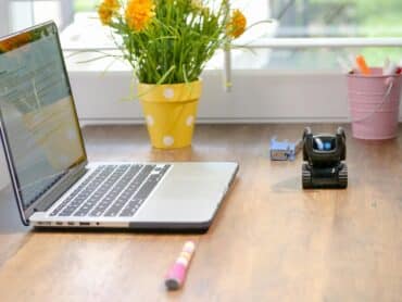 Open laptop on a wooden desk with a yellow polka-dot flower pot in the middle, a pink pen bucket on the right, and a small robot toy nearby.