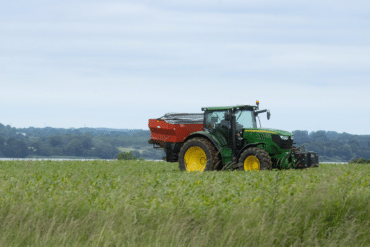 Green John Deere tractor with yellow wheels pulling a red grain trailer across a grassy field under a cloudy sky.